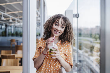 Beautiful woman with bangs having drink in disposable cup at balcony of coffee shop - EBBF03911