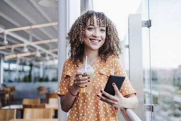 Lächelnde junge Frau mit Mobiltelefon, die wegschaut, während sie auf dem Balkon eines Cafés einen Eistee trinkt - EBBF03913
