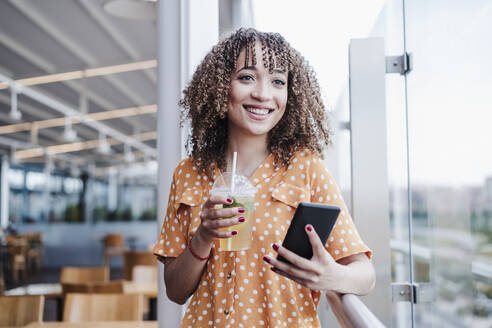 Smiling young woman with mobile phone looking away while having iced tea in balcony at cafe - EBBF03913