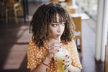 Young woman with curly hair looking away while drinking iced tea in cafe - EBBF03922
