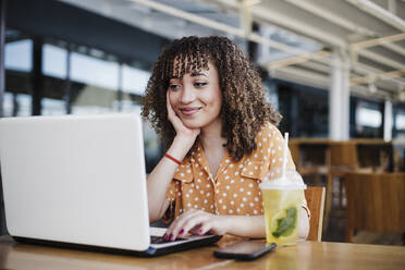 Lächelnde Geschäftsfrau mit Hand am Kinn, die in einem Café einen Laptop benutzt - EBBF03923
