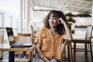 Smiling businesswoman with head in hand day dreaming while sitting by laptop in coffee shop - EBBF03930