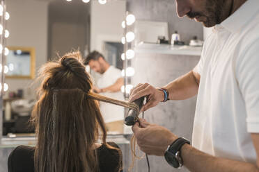 Ein männlicher Friseur bearbeitet das Haar einer Kundin im Salon mit einem Glätteisen - JRVF01003