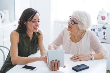 Ältere Frau im Gespräch mit ihrer Enkelin, die ein digitales Tablet benutzt, während sie am Tisch sitzt - OIPF00905
