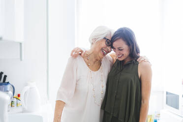 Cheerful grandmother and granddaughter standing with arm around in kitchen at home - OIPF00906