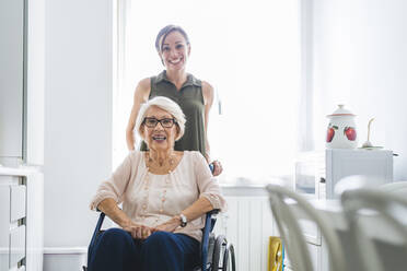 Lächelnde Frau im mittleren Erwachsenenalter mit behinderter Großmutter, die auf einem Stuhl in der Küche zu Hause sitzt - OIPF00924