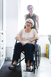 Cheerful disabled senior woman sitting on wheelchair by granddaughter standing in kitchen at home - OIPF00925