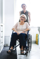 Fröhliche, behinderte Seniorin im Rollstuhl sitzend, neben ihrer Enkelin in der heimischen Küche stehend - OIPF00925