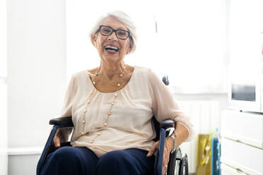 Fröhliche behinderte Seniorin mit Brille, die zu Hause im Rollstuhl sitzt - OIPF00926