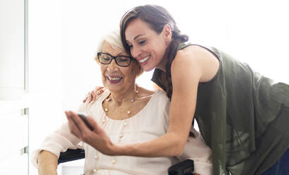Lächelnde Frau, die ein Selfie mit ihrer behinderten Großmutter über ihr Smartphone zu Hause macht - OIPF00928