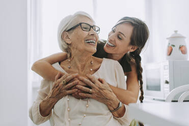 Cheerful woman hugging grandmother sitting in kitchen - OIPF00930