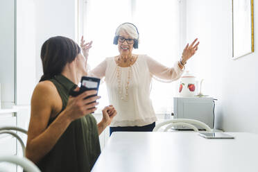 Mittlere erwachsene Frau sitzt mit Smartphone und schaut ihrer Großmutter beim Tanzen zu, während sie zu Hause über Kopfhörer Musik hört - OIPF00942