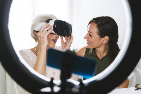 Lächelnde Frau, die ihre Großmutter in einem Virtual-Reality-Simulator betrachtet, während sie zu Hause einen Vlog führt - OIPF00950