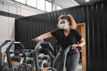 Active woman wearing protective face mask while cycling in gym during COVID-19 - SNF01484