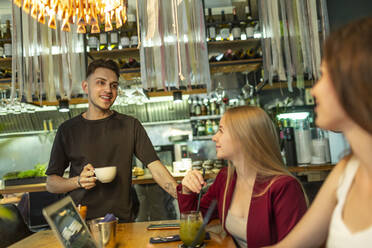 Lächelnder junger Mann mit Kaffeetasse im Gespräch mit Freundinnen am Tisch in einer Bar - VGF00391