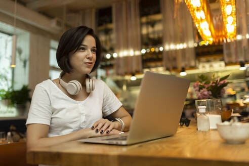 Lächelnde junge Freiberuflerin sitzt mit Laptop im Restaurant und schaut weg - VGF00404