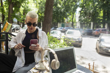 Mittlere erwachsene Frau mit Sonnenbrille, die ein Smartphone benutzt, während sie in einem Café sitzt - VGF00405