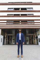 Young businessman looking up while standing on footpath in front of office building - AFVF08886