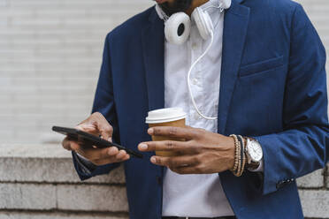 Businessman holding disposable cup while using mobile phone in front of wall - AFVF08890