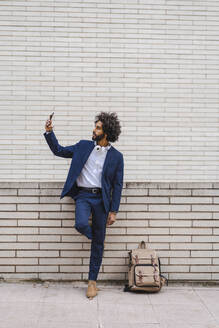 Geschäftsmann, der ein Selfie mit seinem Mobiltelefon macht, während er mit seinem Rucksack auf dem Fußweg steht - AFVF08892