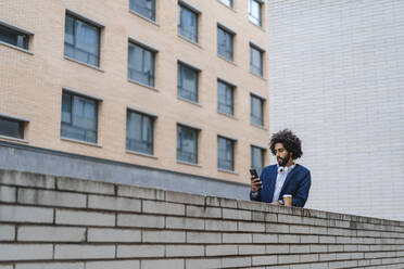 Männlicher Unternehmer, der ein Mobiltelefon benutzt, während er sich auf eine Stützmauer stützt - AFVF08895