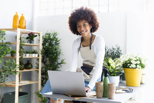 Smiling female shop owner with laptop sitting on table at plant store - GIOF12930