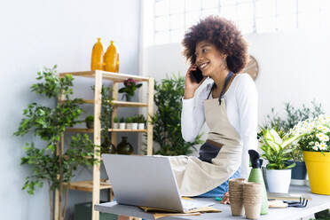 Female florist talking on mobile phone while sitting by laptop on table - GIOF12932