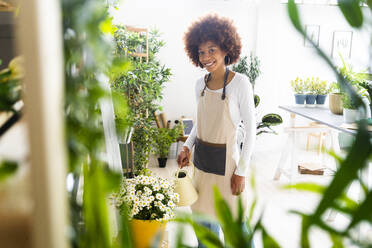 Female florist smiling while watering plant at shop - GIOF12952