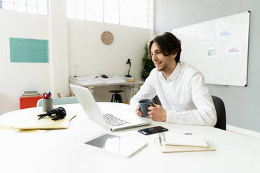 Männlicher Unternehmer mit Kaffeetasse und Laptop bei der Arbeit im Kreativbüro - GIOF12982