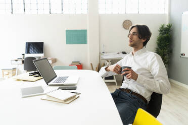 Nachdenklicher Geschäftsmann mit Kaffeetasse am Schreibtisch sitzend im Büro - GIOF12983