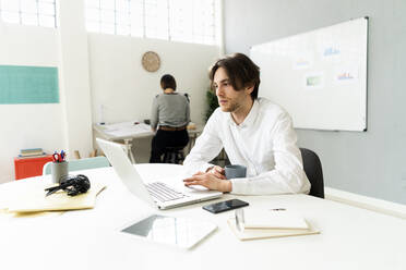 Männlicher Unternehmer, der einen Laptop benutzt, während er mit einem Kollegen im Hintergrund im Büro sitzt - GIOF12984