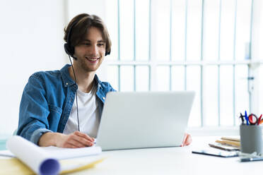 Lächelnder Geschäftsmann mit Headset, der einen Laptop bei der Arbeit im Büro benutzt - GIOF13006