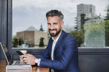 Geschäftsmann mit Mobiltelefon und Laptop, der in einer Cafeteria sitzt und lächelt - EBBF03942