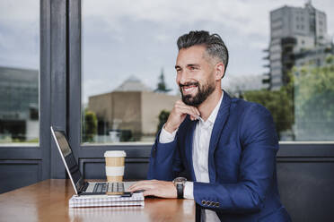 Thoughtful businessman with hand on chin sitting by laptop at cafeteria - EBBF03943