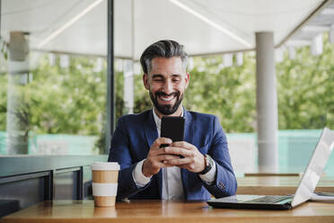 Male entrepreneur with laptop using mobile phone while sitting at cafeteria - EBBF03945