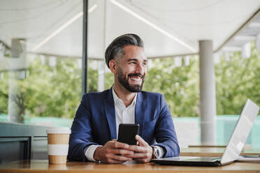 Smiling male entrepreneur with mobile phone and laptop sitting at cafeteria - EBBF03947