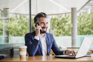 Smiling businessman with laptop talking on smart phone at cafeteria - EBBF03949