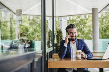 Businessman with disposable cup and laptop talking on mobile phone by window at cafe - EBBF03950