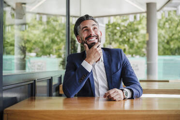 Thoughtful businessman with hand on chin smiling at cafeteria - EBBF03953