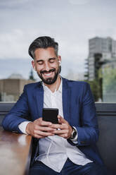 Male entrepreneur with in-ear headphones using mobile phone by table - EBBF03967