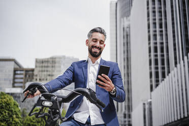 Businessman using mobile phone while standing with rented electric bicycle in city - EBBF03973