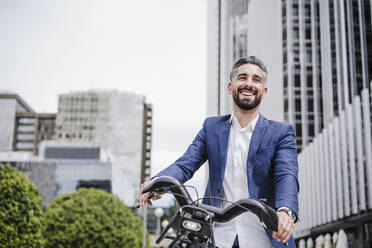 Smiling male entrepreneur standing with rented electric bicycle in city - EBBF03975