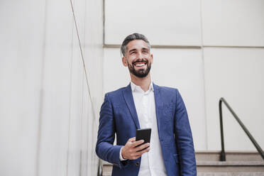 Male entrepreneur with smart phone smiling while standing by wall - EBBF03980