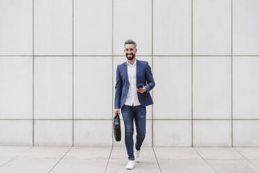 Smiling businessman with briefcase and mobile phone walking on footpath - EBBF03985