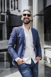 Cheerful businessman with hands in pockets leaning on glass railing - EBBF04012