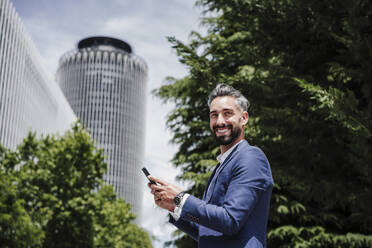 Lächelnder Geschäftsmann mit Mobiltelefon in der Stadt an einem sonnigen Tag - EBBF04015