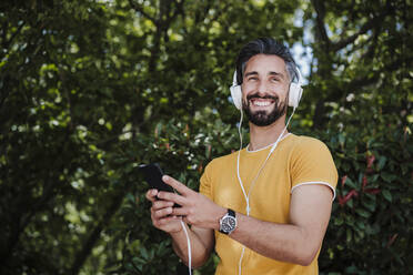 Lächelnder Mann mit Kopfhörern und Mobiltelefon im Park - EBBF04026