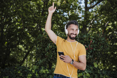 Glücklicher Mann mit erhobener Hand, der im Park über Kopfhörer Musik hört - EBBF04027