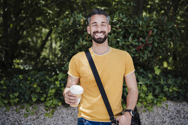 Mid adult man with crossbody bag and disposable cup at park - EBBF04028