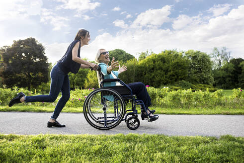 Glückliche Enkelin läuft mit Großmutter im Rollstuhl sitzend im Park - OIPF00957